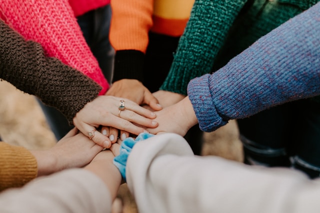 Das Bild steht in Zusammenhang mit Mediation und Konfliktlösung durch die Kanzlei Ponschab & Partner und zeigt folgendes: Eine Gruppe von Menschen, die sich in Teamentwicklung engagieren, steht in einem Kreis und legt in der Mitte ihre Hände übereinander. Sie tragen bunte Pullover – rosa, orange, grün, braun und blau – vor einem unscharfen Hintergrund. Bei Fragen zu Konflikten und Konfliktlösung wenden Sie sich bitte an uns unter info@ponschab-partner.com