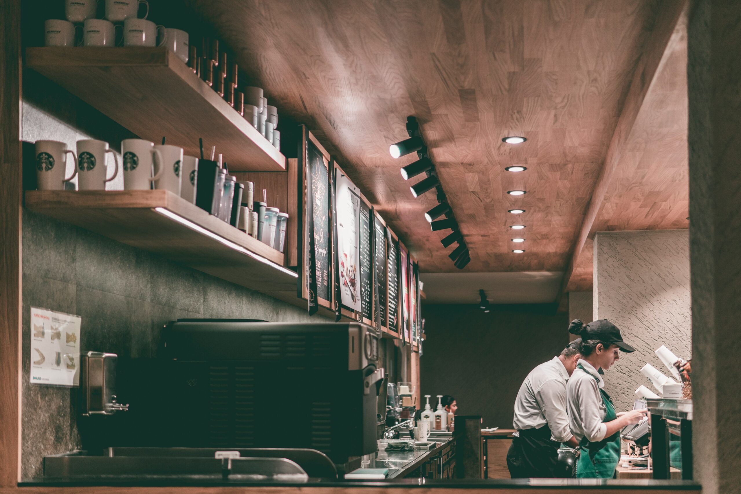 Das Bild steht in Zusammenhang mit Mediation und Konfliktlösung durch die Kanzlei Ponschab & Partner und zeigt folgendes: Zwei Angestellte in Uniform arbeiten hinter dem Tresen eines Coffeeshops, der zu einem Franchise-Unternehmen gehört. Zu sehen sind Regale mit Markenbechern, ein Menübrett und Geräte. Warme Beleuchtung und hölzerne Akzente unterstreichen die einladende Atmosphäre. Das Bild steht im Zusammenhang mit Konfliktlösungen im Franchise. Bei Fragen zu Konflikten und Konfliktlösung wenden Sie sich bitte an uns unter info@ponschab-partner.com