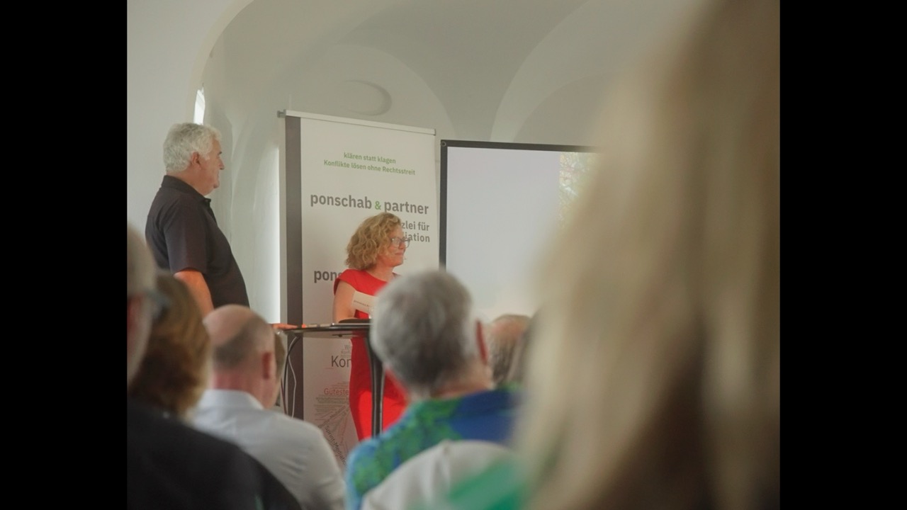 Das Bild steht in Zusammenhang mit Mediation und Konfliktlösung durch die Kanzlei Ponschab & Partner und zeigt folgendes: Ein Mann und eine Frau stehen im vorderen Teil eines Raumes und halten einen Vortrag vor einem Publikum. Die Frau sitzt an einem Tisch und der Mann steht neben ihr. Hinter ihnen sind ein Transparent und eine Projektionsfläche zu sehen. Bei Fragen zu Konflikten und Konfliktlösung wenden Sie sich bitte an uns unter info@ponschab-partner.com