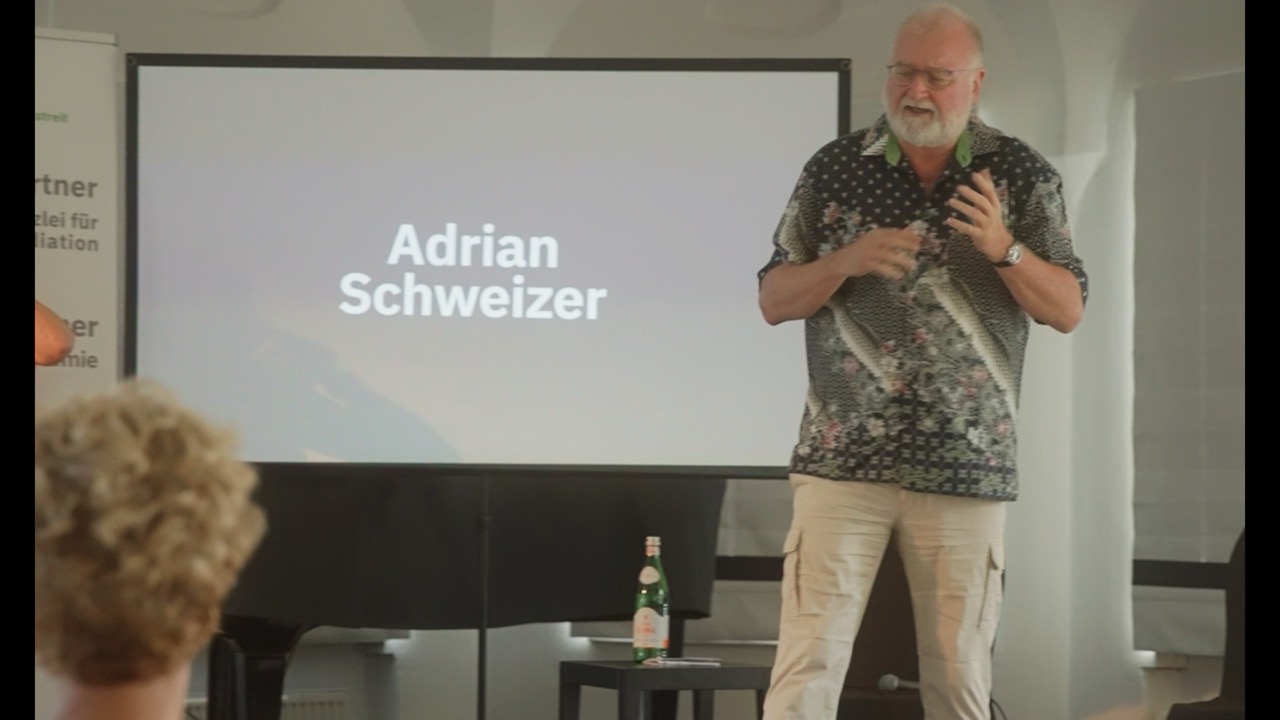 Das Bild steht in Zusammenhang mit Mediation und Konfliktlösung durch die Kanzlei Ponschab & Partner und zeigt folgendes: Ein Mann steht und spricht vor einem Bildschirm, auf dem der Name Adrian Schweizer steht. Auf einem kleinen Tisch in der Nähe steht eine Wasserflasche, und im Vordergrund ist ein Teil des Publikums zu sehen. Bei Fragen zu Konflikten und Konfliktlösung wenden Sie sich bitte an uns unter info@ponschab-partner.com
