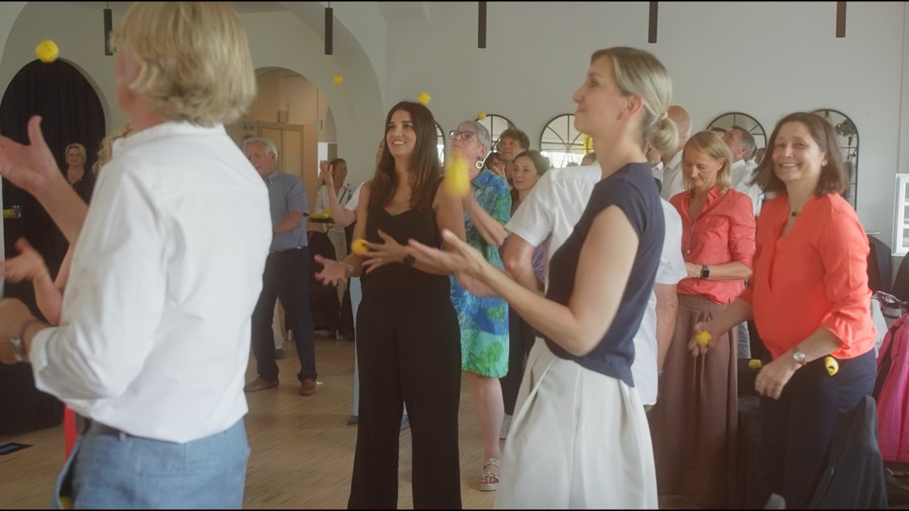 Das Bild steht in Zusammenhang mit Mediation und Konfliktlösung durch die Kanzlei Ponschab & Partner und zeigt folgendes: Eine Gruppe Erwachsener steht in einem Haus und jongliert mit gelben Bällen. Sie wirken engagiert und konzentriert, einige lächeln. Die Umgebung hat gewölbte Türöffnungen und runde Spiegel an der Wand. Bei Fragen zu Konflikten und Konfliktlösung wenden Sie sich bitte an uns unter info@ponschab-partner.com