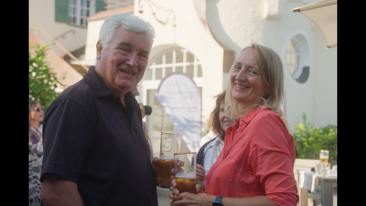 Das Bild steht in Zusammenhang mit Mediation und Konfliktlösung durch die Kanzlei Ponschab & Partner und zeigt folgendes: Ein Mann und eine Frau stehen im Freien, lächeln in die Kamera und halten Biergläser in der Hand. Im Hintergrund sind an einem sonnigen Tag andere Menschen und ein weißes Gebäude mit Fenstern zu sehen. Bei Fragen zu Konflikten und Konfliktlösung wenden Sie sich bitte an uns unter info@ponschab-partner.com