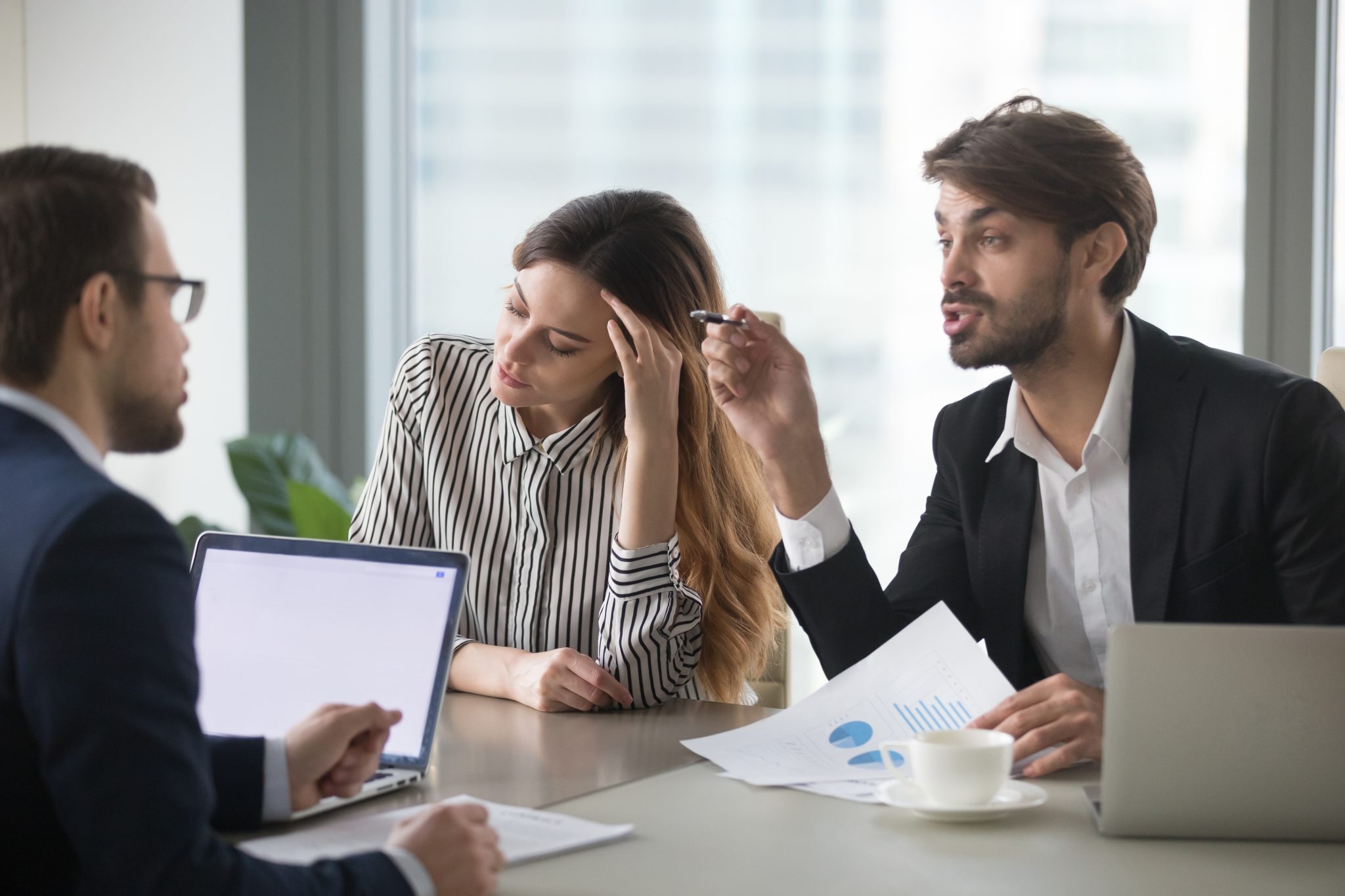 Das Bild steht in Zusammenhang mit Mediation und Konfliktlösung durch die Kanzlei Ponschab & Partner und zeigt folgendes: Drei Personen sitzen an einem Tisch in einem Büro. Ein Mann gestikuliert beim Sprechen, eine Frau schaut frustriert mit der Hand auf dem Kopf, und ein anderer Mann hört zu. Laptops, Dokumente und eine Kaffeetasse stehen auf dem Tisch. Bei Fragen zu Konflikten und Konfliktlösung wenden Sie sich bitte an uns unter info@ponschab-partner.com